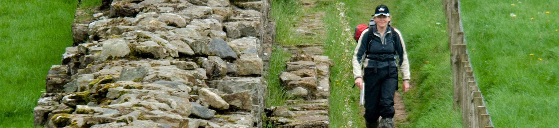 A hiker with a map walks along a trail beside a ruined Roman wall.