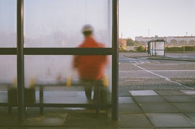 The back view of a person sitting at a bus stop wearing a bright orange jacket which can be seen through the wet frosted glass.
