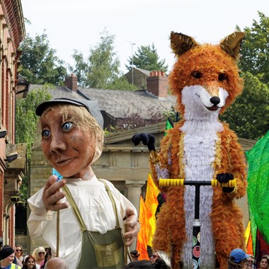 A large fox puppet riding a scooter alongside a humanoid puppet in working overalls at a street carnival