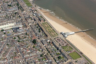 Aerial view of a seaside town, bordering a beach with a strip of gardens between the town and the beach. The sea fills the top right of the picture.