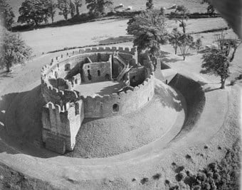 A low altitude aerial photograph of the remains of a circular, crenellated, stone-walled castle, set on a grassy mound surrounded by a ditch and bank.