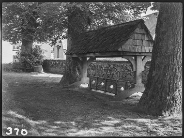 Black and white photograph of village stocks with a pitched timber roof, set between two trees and adjacent to a stone wall. The stocks have four circular leg holes.