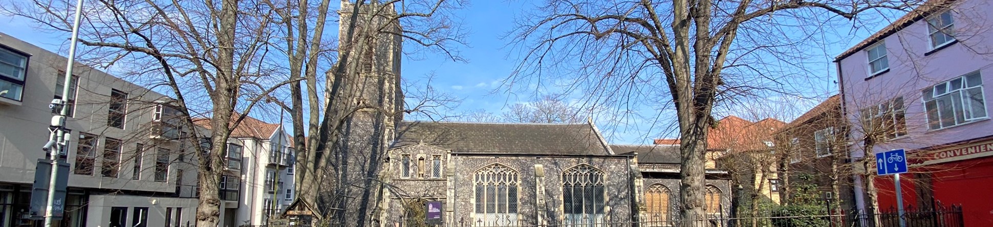 A view from across the street showing the south side of St Margaret de Westwick Church bathed in sunlight. The building is built of grey flint with brick dressings and has plain glass windows. The tower can be seen on the left hand side. Through the wrought iron railings of the churchyard, trees, grass and rows of daffodils can be seen.