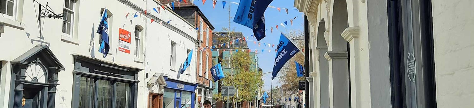 A man walking in a high street lined with historic shops. There is coloured bunting strung across the street.