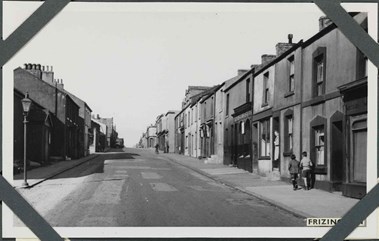 Black and white photograph of an urban street scene. The view looks along a straight road that has a gentle incline. The road is flanked on either side by mainly two-storey houses and commercial premises that open directly onto the pavement. At the left of the photograph, a street lamp stands at the edge of the pavement. On the opposite side, two boys walk side-by-side away from the camera, approaching a woman standing on a doorstep. A label applied to the bottom-right of the photograph reads: 'Frizington', part of which is obscured by one of four corner photograph album mounts.