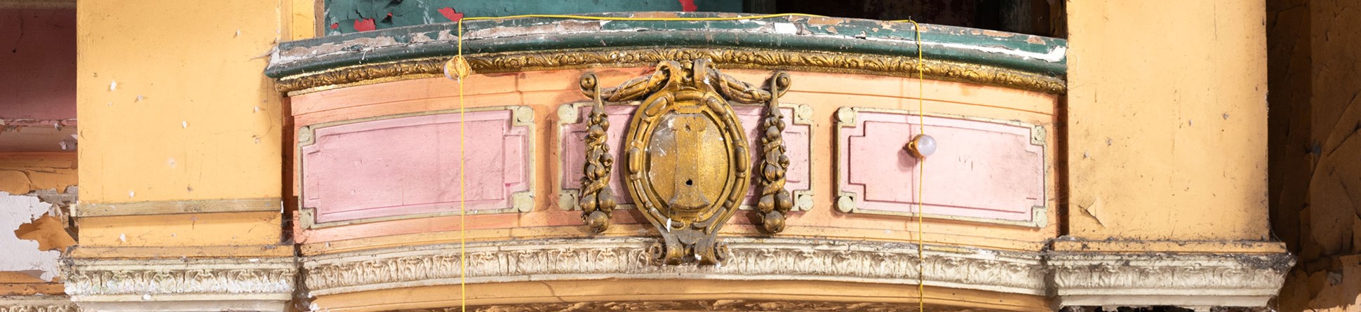 Detail of an ornate but derelict theatre interior, a balcony front with decorative moulding, flanked by columns.