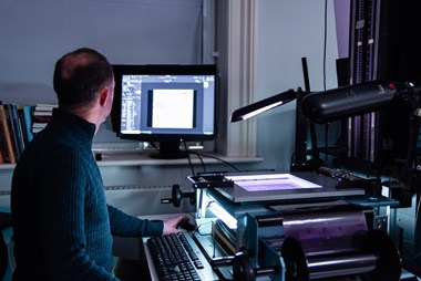 A man at work digitising photographic film. The man sits at a desk that supports a keyboard and equipment set up to digitally capture images from photographs, in this case a roll of photographic film negatives. Angled lights illuminate the negative. The member of staff looks towards a monitor.