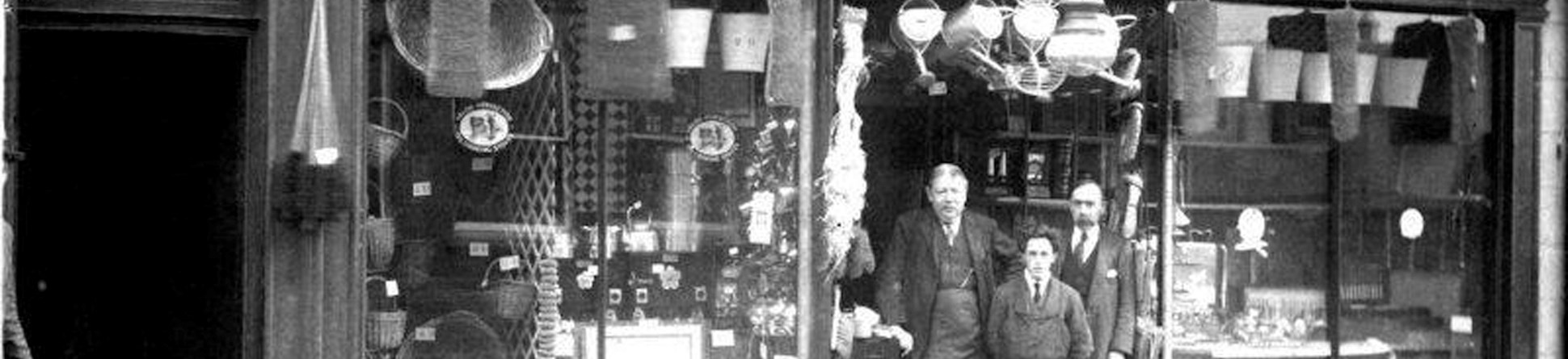 A black and white photograph shows three men wearing shirt, tie, and apron, standing in the doorway of an ironmonger's shop. The large windows either side of the doorway are filled with wares and a tin bath, watering cans and other objects hang above the door.