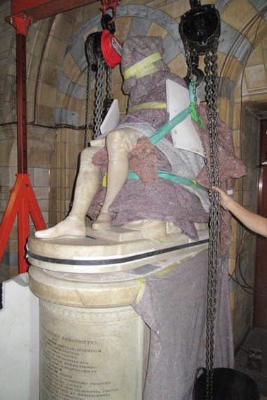 A stone statue of a seated figure covered in blankets and foam padding is lifted by two winches.