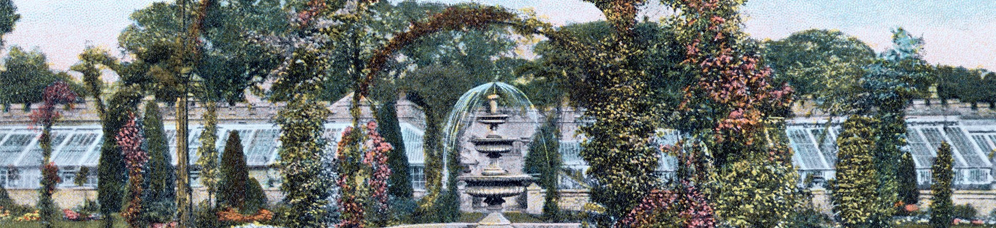 A colourised historic postcard shows a central fountain with arches in the foreground and flower borders either side of a grass path. Glass houses can be seen in the background.