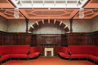 Red velvet benches line wood-panelled walls. Above the panelling, the walls are covered in ceramic tiling. In the centre of the image is an arched recess with a marble fireplace and wood-panelled mantelpiece.