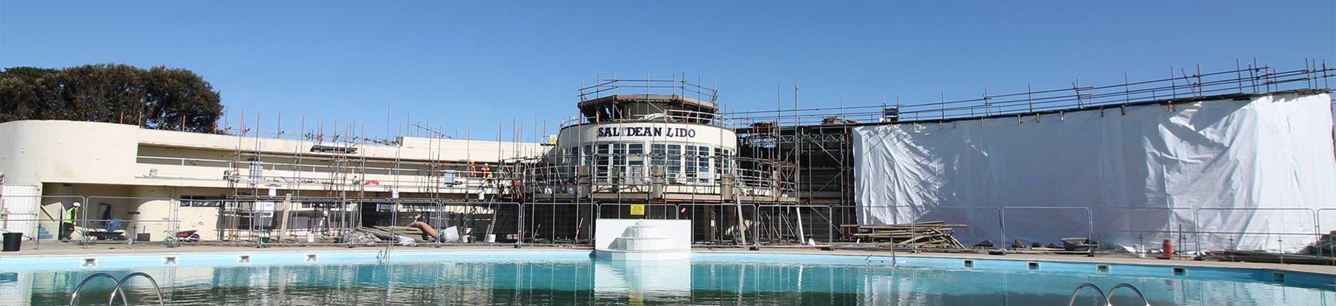 Wide cream-coloured building with a swimming pool in front has scaffolding on it.