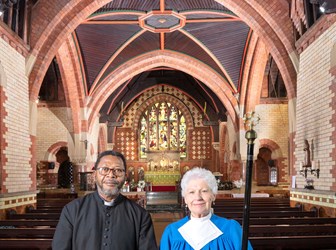 A priest and colleague stand in the nave of a church, the altar and stained glass windows behind them.