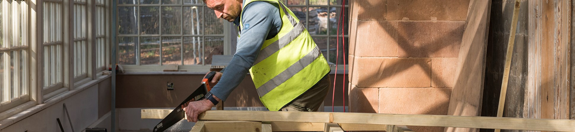 Man in a conservatory undergoing renovation. He is wearing a high-vis waistcoat and sawing a length of wood balanced on a frame made from the same wood.