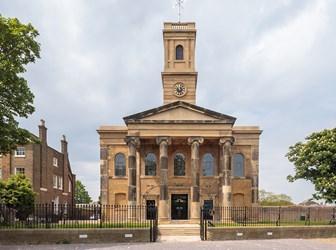 Front view of a sandstone church with darkened features, columns, pediment and square tower. 