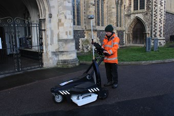 Man in high-vis jacket operating ground-penetrating survey equipment in the grounds of a church.