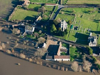 Birds-eye view of a village with bare trees and building foundations. Greyish-brown water has flooded the bottom half of the image. 