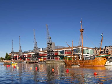 A photograph of four large metal cranes on a dockside in front of an industrial building. A reconstructed historic wooden ship is moored in front of them.