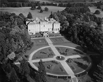 An aerial photograph taken at low altitude showing the garden front of a country house. Clusters of trees flank the house and surround the gardens. 