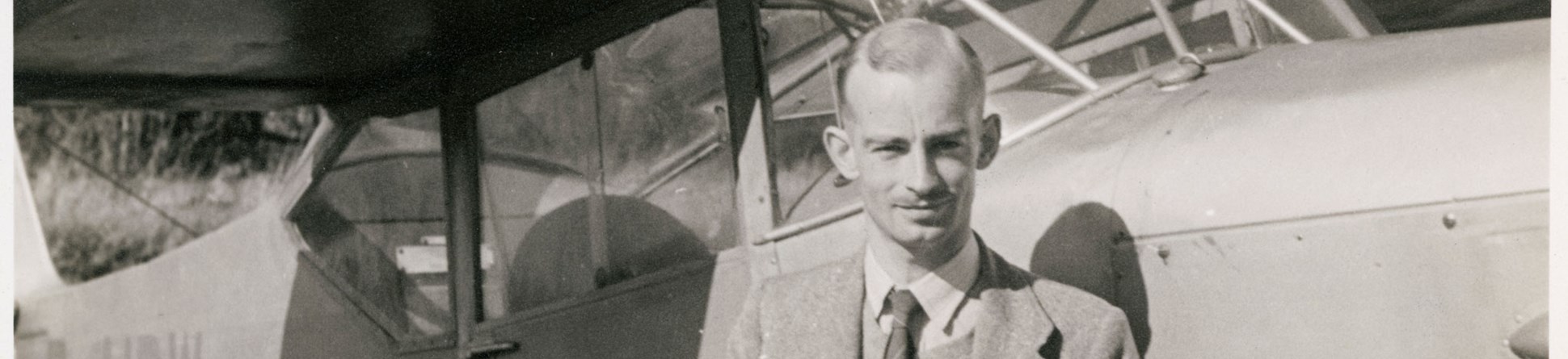 A photograph of Harold Wingham as a young man, posed in front of light aircraft. Pinned to his tie is a badge or medallion featuring a winged motif.