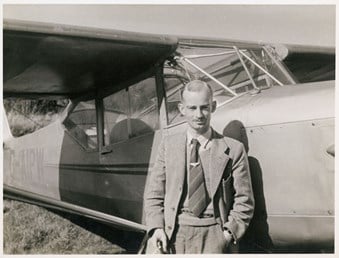 A photograph of Harold Wingham as a young man, posed in front of light aircraft. Pinned to his tie is a badge or medallion featuring a winged motif.