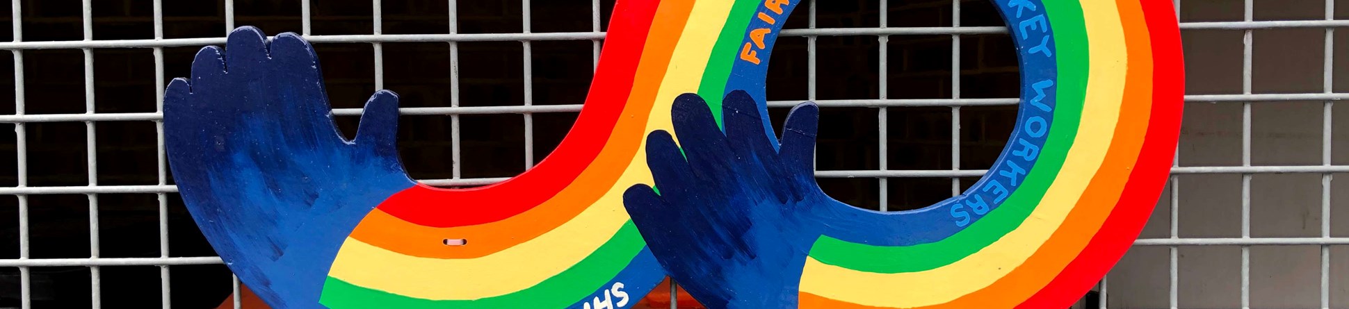 Rainbow colours with hands at each. Words 'Support Our NHS, Fair Wages for Key Workers' written on the blue band of the rainbow. The graphic is attached to a metal fence.