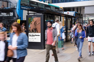 A bus pulled into a bus stop, on a busy high street. A clear channel digital screen at the front of the bus stop shows a picture of a Shakespeare Playhouse North. The title reads 'Bus Stop Stories'