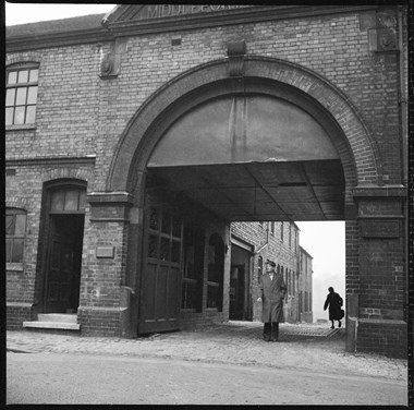 View of brick industrial building with large arched entrance. A man in a mackintosh and cap stands in the entrance. The silhouette of a woman walking away from camera is visible on the far side of the entrance.